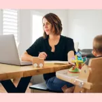 Woman working from home in front of computer with child in high chair - work life balance